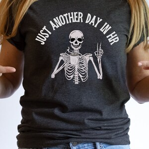 May include: A dark gray t-shirt with the text "Just another day in HR" and a skeleton holding a coffee cup and giving the middle finger.