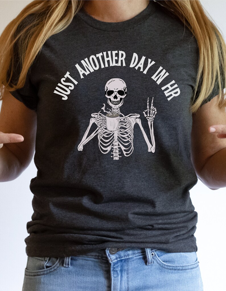 May include: A dark gray t-shirt with the text "Just another day in HR" and a skeleton holding a coffee cup and giving the middle finger.
