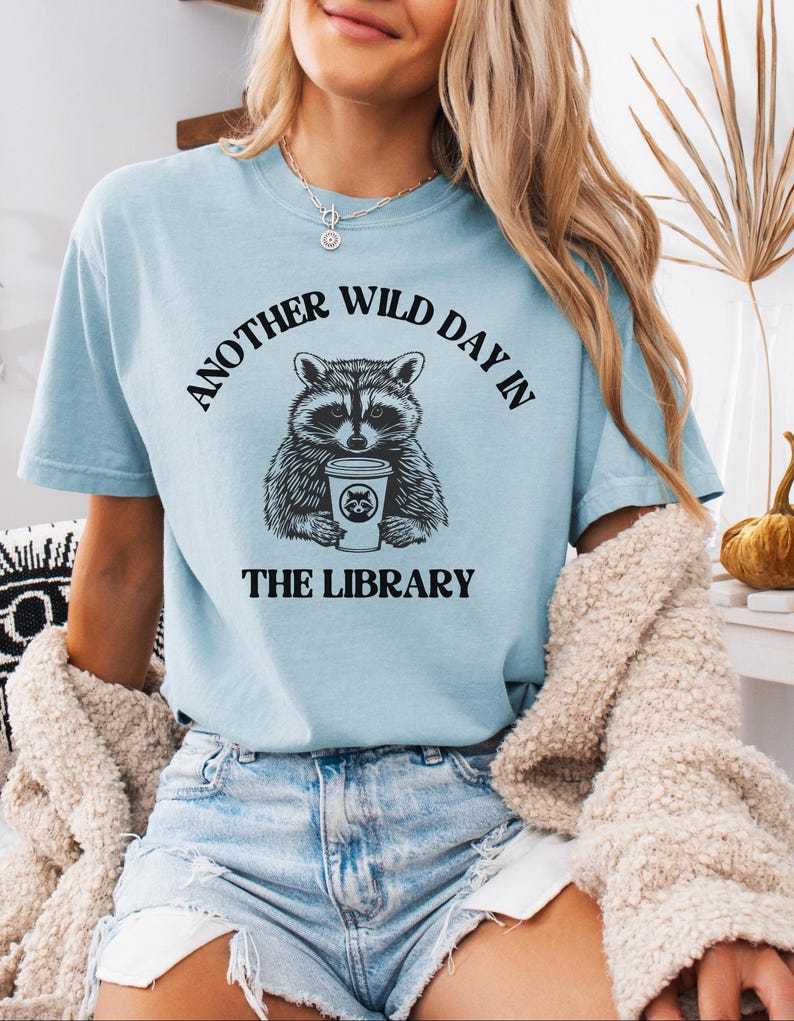 Peut inclure: T-shirt bleu clair avec le texte "ANOTHER WILD DAY IN THE LIBRARY" au-dessus et en dessous d'un dessin de raton laveur tenant une tasse de caf&eacute;. Le dessin du raton laveur est en noir et blanc. Le t-shirt est en mati&egrave;re souple.