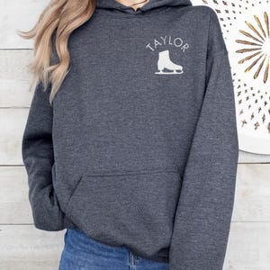 K&ouml;nnte beinhalten: Ein dunkelgrauer Kapuzenpullover mit dem Wort "TAYLOR" und einer wei&szlig;en Silhouette einer Schlittschuhs auf der Vorderseite.
