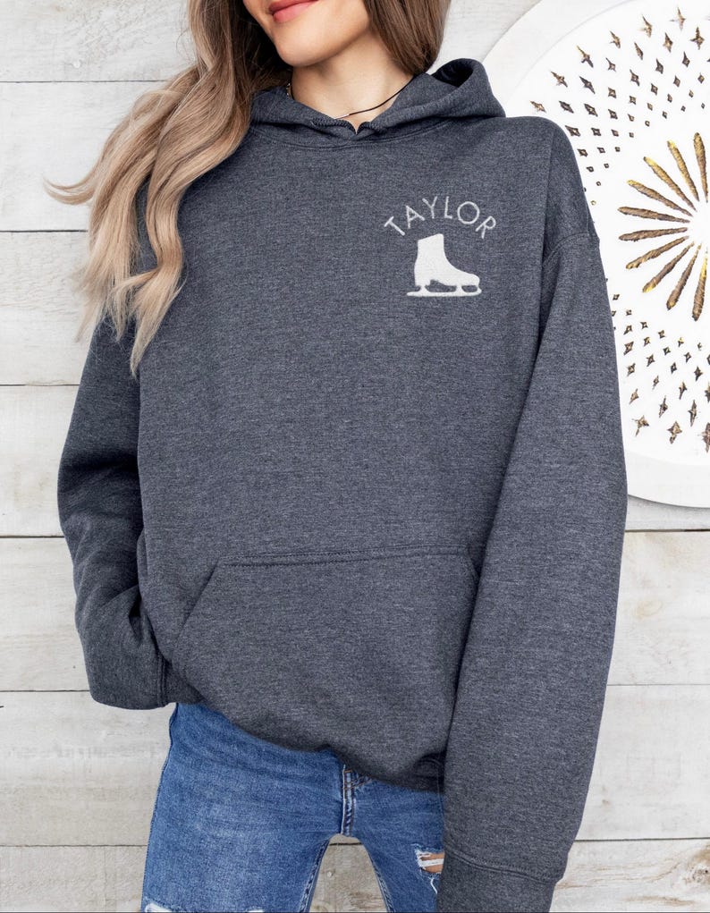 K&ouml;nnte beinhalten: Ein dunkelgrauer Kapuzenpullover mit dem Wort "TAYLOR" und einer wei&szlig;en Silhouette einer Schlittschuhs auf der Vorderseite.