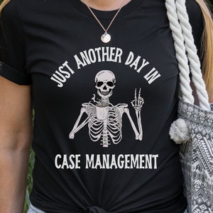 May include: Black t-shirt with a white skeleton holding a coffee cup and the text "Just another day in case management".