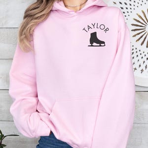 K&ouml;nnte beinhalten: Ein rosa Hoodie mit einem schwarzen Schlittschuh-Motiv und dem Wort "TAYLOR" auf der Vorderseite.