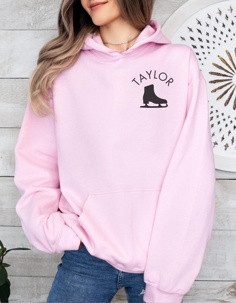 K&ouml;nnte beinhalten: Ein rosa Hoodie mit einem schwarzen Schlittschuh-Motiv und dem Wort "TAYLOR" auf der Vorderseite.