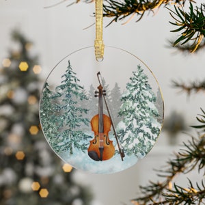 May include: A round glass Christmas bauble featuring a watercolour illustration of a violin in a snowy woodland. The violin is brown with a light brown bow. The trees are green with white snow on the branches.