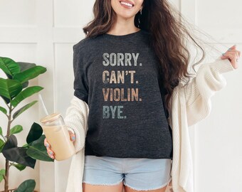 May include: A dark gray t-shirt with the text "SORRY. CAN'T. VIOLIN. BYE." in a vintage style font. The text is in shades of blue, pink, and yellow.