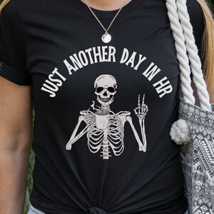 May include: Black t-shirt with a white graphic of a skeleton holding a coffee cup and the text "Just another day in HR".