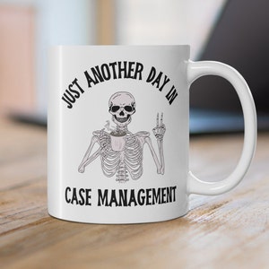 May include: White ceramic mug with a black graphic of a skeleton holding a coffee cup and making a peace sign. The text reads "JUST ANOTHER DAY IN CASE MANAGEMENT".