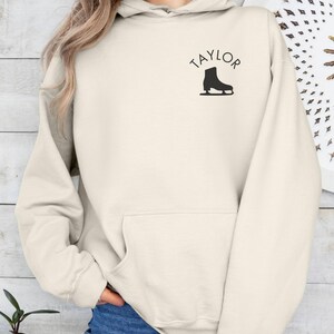 K&ouml;nnte beinhalten: Ein cremefarbener Hoodie mit einer gro&szlig;en Tasche auf der Vorderseite. Der Hoodie hat das Wort "TAYLOR" in schwarzen Buchstaben &uuml;ber einer schwarzen Silhouette einer Schlittschuhs gedruckt.