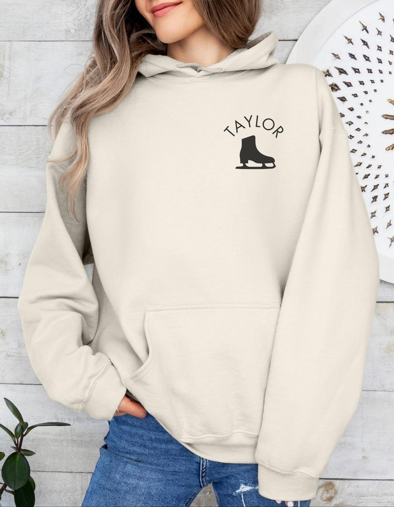 K&ouml;nnte beinhalten: Ein cremefarbener Hoodie mit einer gro&szlig;en Tasche auf der Vorderseite. Der Hoodie hat das Wort "TAYLOR" in schwarzen Buchstaben &uuml;ber einer schwarzen Silhouette einer Schlittschuhs gedruckt.