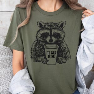 May include: An olive green t-shirt with a black and white raccoon illustration holding a coffee cup. The cup has the text "IT'S WILD IN HR". The shirt has a crew neck and short sleeves.