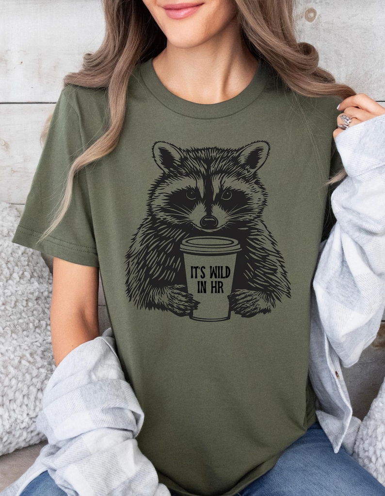 May include: An olive green t-shirt with a black and white raccoon illustration holding a coffee cup. The cup has the text "IT'S WILD IN HR". The shirt has a crew neck and short sleeves.