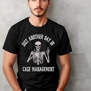 May include: Black t-shirt with a white graphic of a skeleton holding a coffee cup and giving the peace sign. The text reads "Just another day in case management."