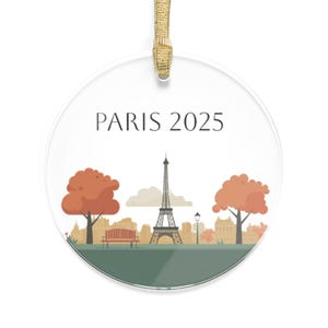 May include: A round, clear ornament with a white background and a green bottom. The ornament features a stylized illustration of the Eiffel Tower in Paris, France, with two trees on either side. The text "PARIS 2025" is printed in black at the top of the ornament.