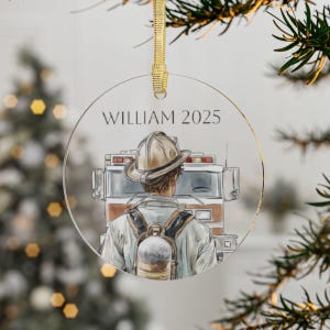 May include: A clear, round Christmas ornament with a watercolor illustration of a firefighter in front of a fire truck. The ornament has the name "WILLIAM 2025" printed on it and is suspended by a gold ribbon.