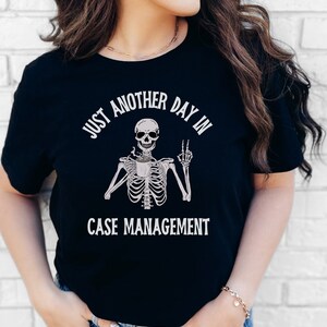 May include: Black t-shirt with a skeleton holding a coffee cup and the text "Just another day in case management".