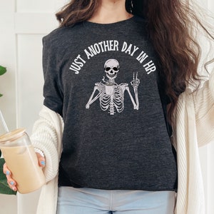 May include: A dark grey t-shirt with a skeleton holding a coffee cup and making a peace sign. The text on the shirt reads "Just another day in HR".