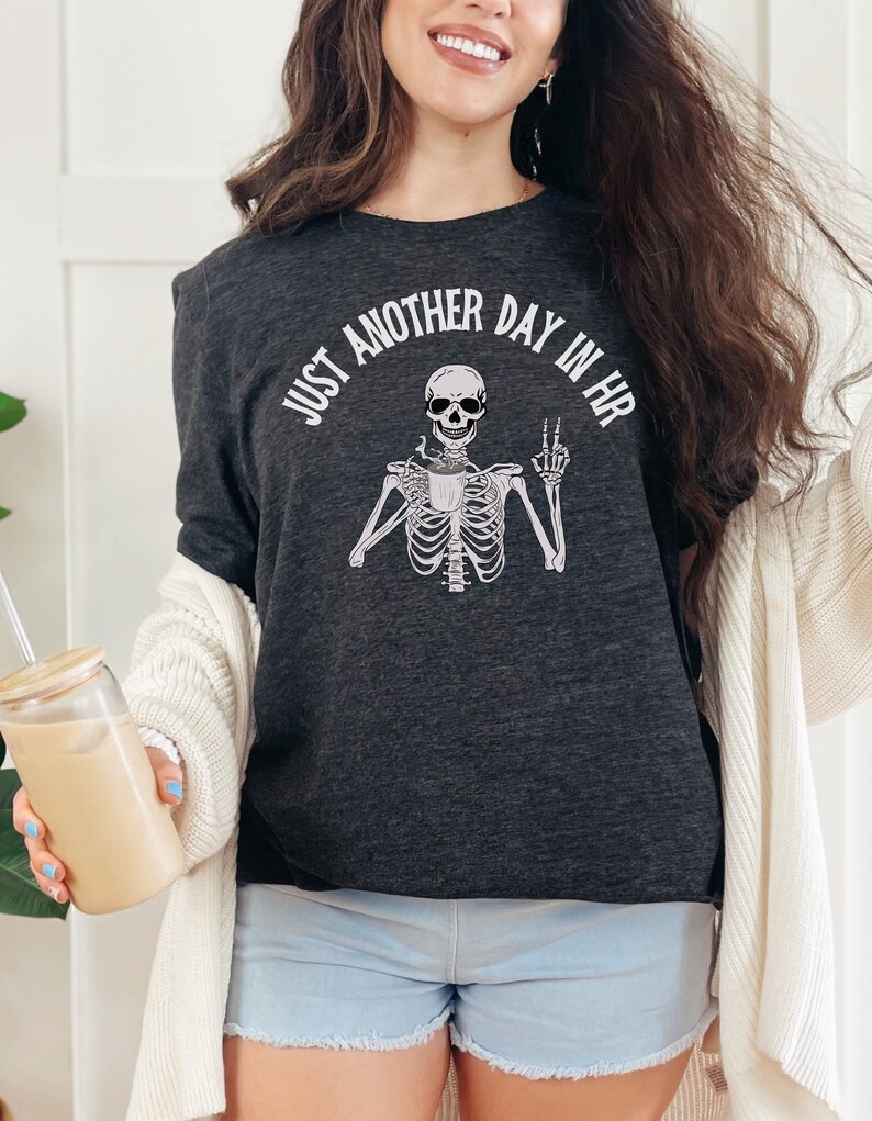 May include: A dark grey t-shirt with a skeleton holding a coffee cup and making a peace sign. The text on the shirt reads "Just another day in HR".