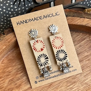 May include: A pair of handmade earrings with a silver lotus flower charm and a rectangular fabric panel with red and black circular patterns. The fabric panel has a white background and the earrings are hanging from a brown card with the text "HANDMADEAHOLIC" printed on it.