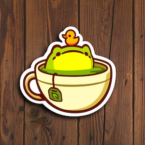 May include: A green frog wearing a yellow rubber ducky sits in a white teacup with a green tea bag. The teacup has a brown rim and a brown handle.