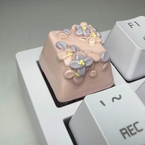 May include: A custom mechanical keyboard keycap in a soft pink color, featuring raised, sculpted light purple flowers with small yellow centers. The keycap is installed on a white keyboard next to a white keycap labeled with the text REC.