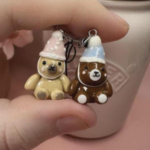 May include: Two small bear charms made of smooth material, one light tan and one dark brown. Each bear wears a tiny hat with white polka dots, one in soft pink and one in light blue. The bears have painted facial features and white paw pads.