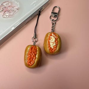 Op de afbeelding: Twee miniatuur gepofte aardappelbedels, één met een zwart koordje en één met een zilveren metalen sleutelhanger. Elke aardappel is bruin met een opening gevuld met oranje bonen, en één heeft ook een klontje witte boter.