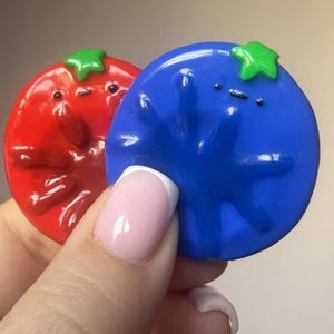 May include: Two circular, glossy, flat-backed charms featuring embossed, stylised faces with simple dot eyes and a small line mouth. One charm is bright red with a green stem top, and the other is vibrant blue with a green stem top.