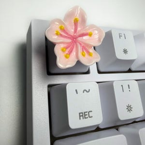 May include: A light pink cherry blossom flower shaped keycap attached to a white mechanical keyboard. The flower features five rounded petals with a darker pink centre and small yellow stamens. The keyboard keys display F1, REC, and symbols.