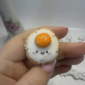 May include: A small, circular, fried egg-shaped charm featuring a glossy white egg white and a raised, smooth, bright orange yolk. The white portion displays a hand-painted, simple black smiling face with two tiny pink dots for cheeks.
