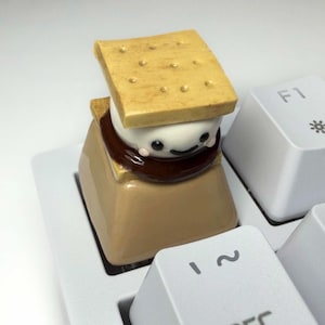May include: A tan mechanical keyboard keycap featuring a s'more design. The s'more has a light brown graham cracker top with small indentations, a white marshmallow center with a small black smiling face, and a dark brown chocolate layer.