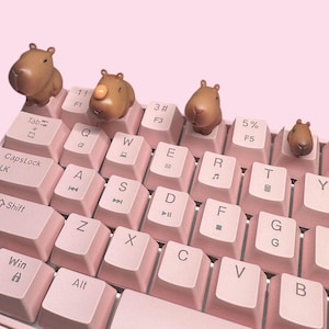 May include: A mechanical keyboard with light pink keycaps featuring standard QWERTY lettering and symbols. Four brown capybara-shaped artisan keycaps are attached to the top row, replacing the Tab, F1, F3, and F5 keys with small animal figures.