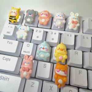 Könnte beinhalten: Eine Sammlung von neun kleinen, bunten, dreidimensionalen tierförmigen Harzfiguren, die auf weißen Computertastaturtasten platziert sind. Die Figuren umfassen eine Biene, einen grauen Koala, einen pfirsichfarbenen Affen, eine weiße Katze, einen weißen Hasen, einen blauen Pinguin, ein gelbes Küken, einen rosa Hasen und einen orangefarbenen Fuchs, alle mit bemalten Gesichtsdetails.