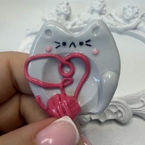 May include: A light gray ceramic cat-shaped trinket dish featuring a hand-painted black face with a simple expression, pink cheeks, and a raised pink yarn ball sculpture attached to the center, creating a decorative and functional small tray.