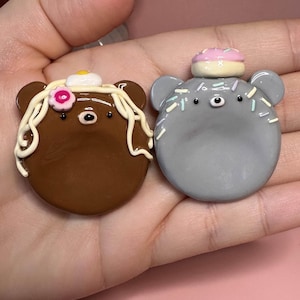 May include: Two small, round, bear-shaped charms. The brown bear features white icing-like swirls, a small pink flower, and a tiny egg on top. The gray bear is decorated with colorful sprinkles and a small pink and cream-colored donut on its head.
