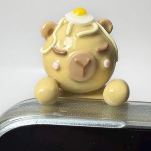 May include: A tan, round, clay-like bear figurine with closed eyes, pink cheeks, and a brown snout. A small white and yellow fried egg sits on top of its head with white noodle-like strands draped over its ears and forehead, attached to a clear phone case.