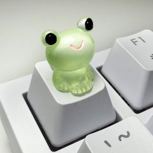 May include: A translucent light green frog figurine attached to the top of a white computer keyboard keycap. The frog has large black eyes and a small pink mouth. The keycap is positioned next to an F1 key on a white mechanical keyboard.