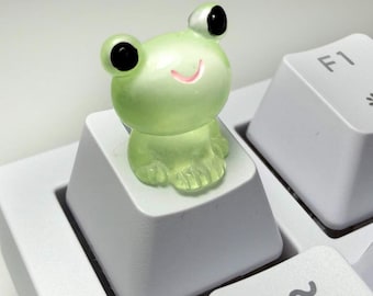 May include: A translucent light green frog figurine attached to the top of a white computer keyboard keycap. The frog has large black eyes and a small pink mouth. The keycap is positioned next to an F1 key on a white mechanical keyboard.