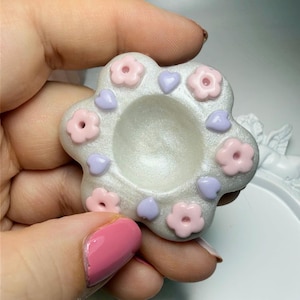 May include: A small, flower-shaped tray with a shimmering white finish. The surface is decorated with alternating light pink flowers and small lavender hearts arranged around a central circular indentation. The object has a smooth texture.