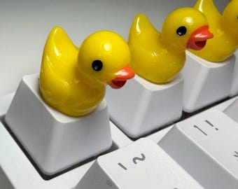 May include: Three bright yellow rubber duck figures attached to white mechanical keyboard keycaps. Each duck has a small orange beak and black eyes. The keyboard keys are white with black lettering, including a key labeled with the text REC.