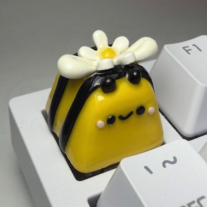 May include: A yellow and black bee-shaped mechanical keyboard keycap featuring a white daisy flower on top. The keycap has a glossy finish, black stripes, and a small painted face with black eyes and a smile, positioned between white keys.