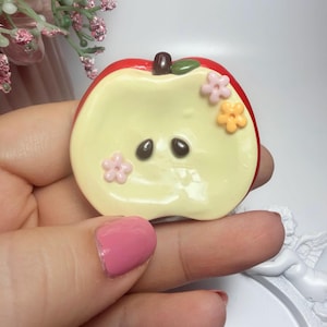 May include: A flat, apple-shaped decorative piece with a red outer edge and a pale yellow centre. It features a small brown stem, a green leaf, two dark brown seeds, and three tiny five-petaled flowers in light pink and orange colours.