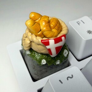 May include: A custom mechanical keyboard keycap featuring a sculpted basket filled with golden-brown croissants. The basket is adorned with a red and white Danish flag emblem and sits atop a textured, dark gray base with green foliage accents.