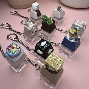 May include: A set of ten keychains featuring miniature food items, each with a different design. The keychains are made of resin and have a clear acrylic base. The food items include a marshmallow, a bear, a plant, a cake, a s'more, a bowl of noodles, and a chocolate bar.