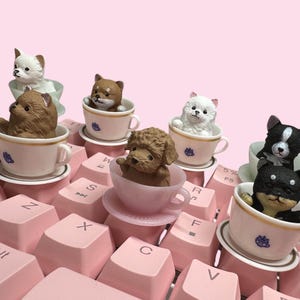 May include: A collection of miniature dog figurines, each seated inside a small teacup and saucer. The dogs include white, brown, and black breeds. The teacups are white with gold rims and small blue crests, resting on a light pink computer keyboard.