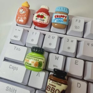 Puede incluir: Cinco teclas de teclado en miniatura con temática de comida sobre un teclado de ordenador blanco. Las teclas presentan diseños de una botella de salsa de pollo, una botella de kétchup, un tarro de crema de cacahuete Skippy, una mermelada de té verde y una salsa barbacoa coreana.