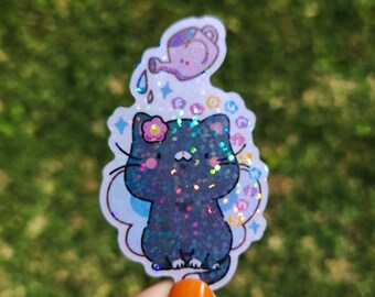 May include: A black cat with a flower on its head and iridescent glitter wings is being watered by a small purple watering can. The sticker is on a white background with a glittery design.