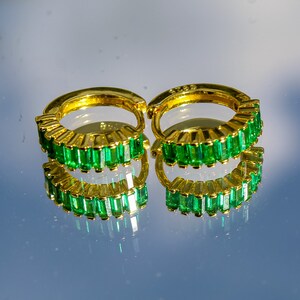 May include: A pair of gold hoop earrings with emerald green baguette-shaped stones set around the hoop.