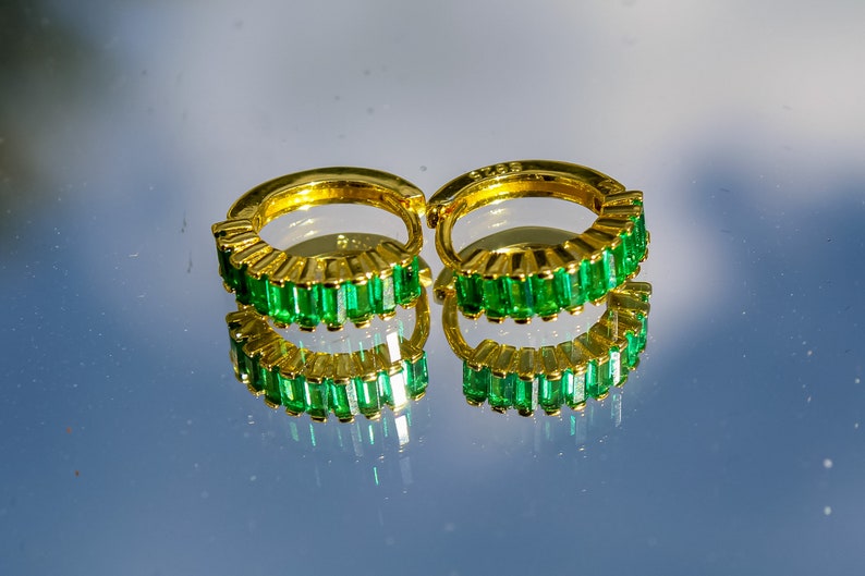 May include: A pair of gold hoop earrings with emerald green baguette-shaped stones set around the hoop.