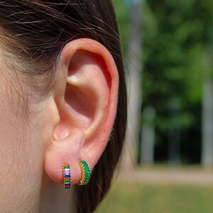 May include: A close-up of a person's ear with two gold hoop earrings. The earring on the left is set with multi-colored stones, while the earring on the right is set with green stones.